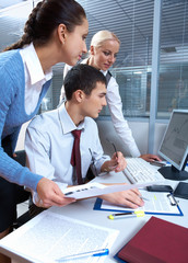 A businessman sitting at table and two women standing over him, all looking at the computer monitor