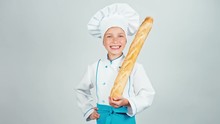 Happy Baker Girl 7-8 Years Child Holds Bread Baguette And Gives You It And Smiling At Camera With Teeth