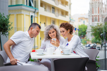 Family day. Elderly loving couple and their adult daughter using smartphone together while sitting in sidewalk cafe.