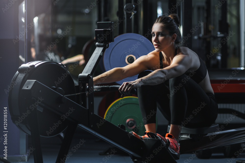 Concept: power, strength, healthy lifestyle, sport. Powerful attractive muscular woman CrossFit trainer do workout on indoor rower at the gym - obrazy, fototapety, plakaty 