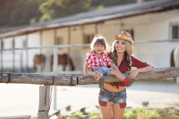 Young woman in cowboy hat,spends time in the countryside in the fresh air in summer with his young daughter,girl,blonde,mother and daughter are dressed in red checkered shirt and shorts,blue denim