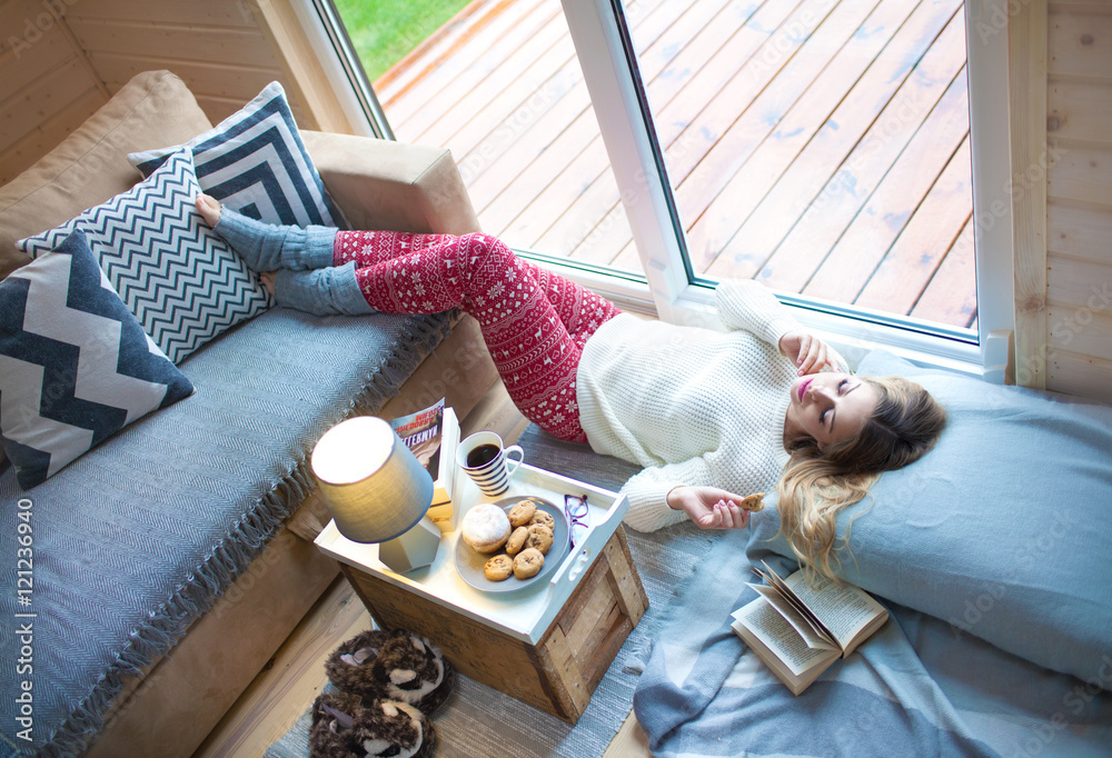 Young beautiful blonde woman with book lying down home by the livingroom window. Lazy cold day off concept - obrazy, fototapety, plakaty 