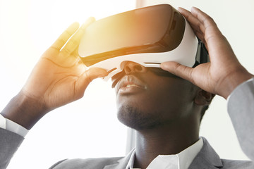 Close up of dark-skinned player dressed in formal wear with virtual reality headset or 3d glasses on his head, playing video game. African man wearing goggles against white studio background