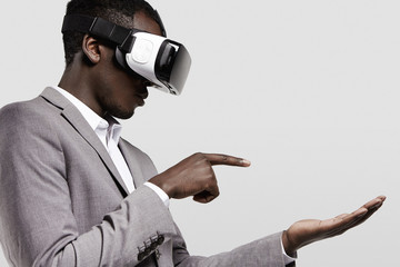 Dark-skinned man in formal wear with virtual reality headset for smart phone on his head, playing video games., holding his hands as if typing message on electronic device, looking concentrated