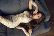 Young sexy woman in white lace evening dress lyuing on gray couch. Top view. Gorgeous glamorous girl with long brunette curly hair, makeup and red lips looking at you