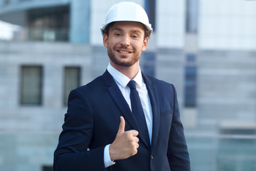 Great work! Handsome young architect showing his thumb up and looking at camera with smile while standing outdoors 