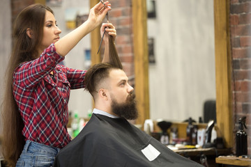 Side view portrait of handsome young bearded caucasian man getting trendy haircut in modern barber shop. Attractive hairstylist girl working, serving client, doing haircut using metal shears and comb