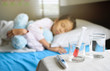 © maemanee - Generic liquid medicine syringe in glass on table in front of out of focus sleeping child for health and illness concept, young Asian girl hugs the blue teddy bear.