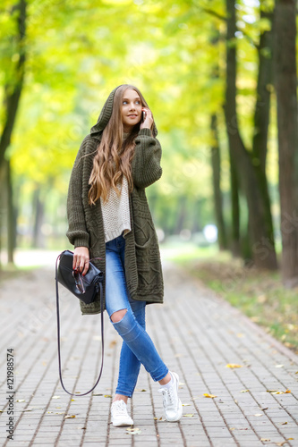 Fotografía Beautiful young woman in green knitted cardigan with a hood walking in autumn park and talking mobile phone