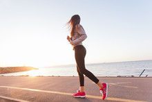 Young Woman Athlete Running At Sunset On Seashore Sidewalk. Girl Runner Training On A Beach