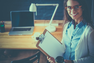 Young woman standing near desk with laptop holding folder and cup of coffee