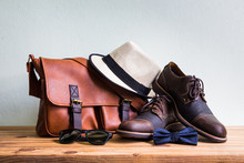 Men's Accessories With Brown Leather Bag, Brown Shoes, Classic Hat, Sunglasses And Blue Bow Tie On Wooden Table Over Wall Background