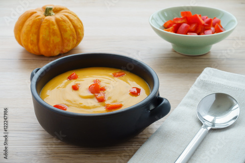 Fotografie, Obraz Pumpkin soup with chopped red peppers in a brown ceramic bowl with handles, a munchkin pumpkin, a bowl with red chopped peppers, a grey napkin and a soup spoon on light brown wooden background