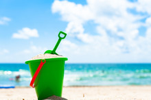 Lone Green Child's Beach Toy Sand Bucket Pail And Shovel With Sea And People Playing In Background