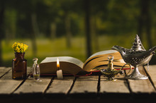 Thick Book Lying Open On Wooden Surface, Small Brown Bottle With Flowers, Aladin Style Lamps And Wax Candle Next To It, Magic Concept Shoot
