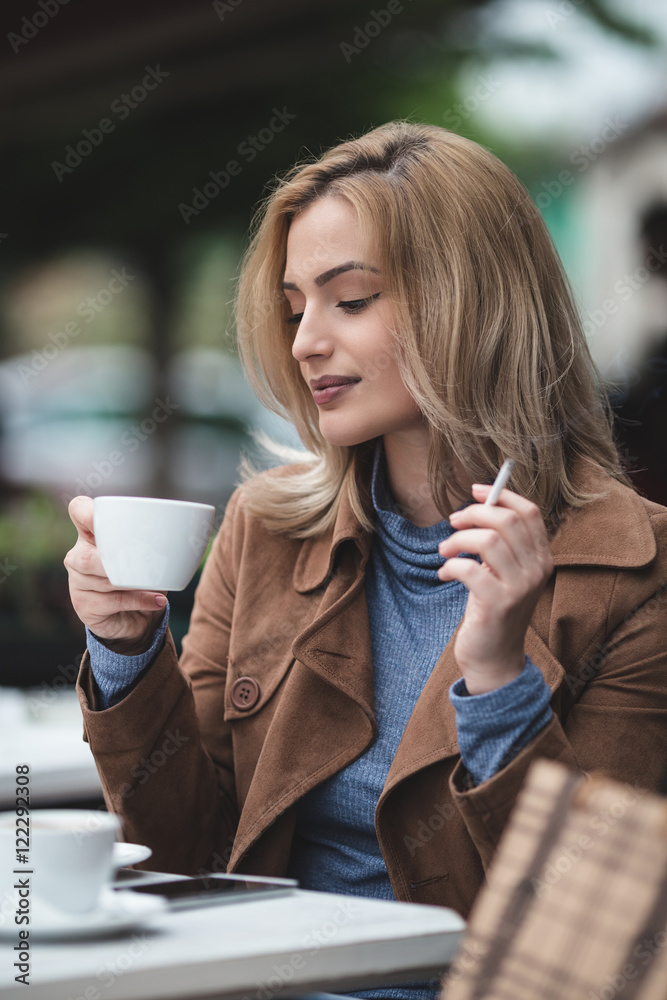 smoking cigarettes and drinking coffee Modern lifestyle. Beautiful young blonde woman enjoying city outdoor. She  siting in cafe bar, drinking coffee and smoking cigarette. Stock Photo