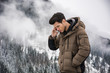 Dark haired handsome young man in winter outerwear using cell phone or smartphone, outdoor at mountain with snowy landscape behind