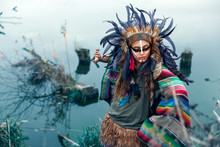 Beautiful Girl Dressed In Native American Indian Clothes, With Paint Face Camouflage And Headdress With Black Feathers And Fur Dancing In Front Of Lake In Summer