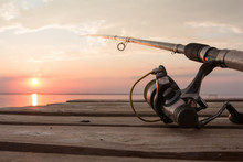 Fishing Reel And Rod Lying On Wooden Pier Over The Sunset Lake