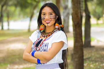 Wall Mural - Beautiful Amazonian woman with indigenous facial paint and white traditional dress posing happily for camera in park environment, forest background