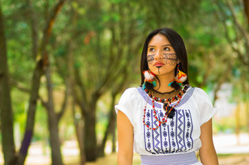 Wall Mural - Beautiful Amazonian woman with indigenous facial paint and white traditional dress posing happily for camera in park environment, forest background