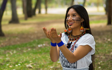 Wall Mural - Beautiful Amazonian woman with indigenous facial paint and white traditional dress posing happily for camera in park environment, forest background