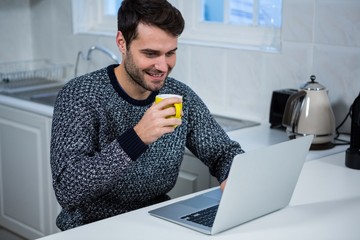 Poster - Man holding coffee mug while using laptop in the kitchen