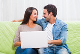 Fototapeta Tulipany - Happy couple sitting on sofa at their home and using laptop.