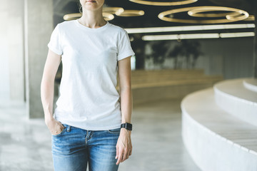 Wall Mural - Girl in white T-shirt and blue jeans standing indoor with her hand in her pocket.On second hand digital gadget smartwatc.Nearby places for sitting with wooden texture. Mock up.