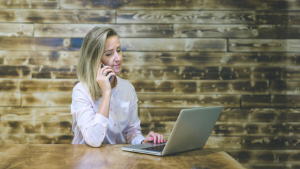 Wall Mural - Young business woman sitting at a table, talking on cellphone and checking e-mail on a laptop. Beautiful girl sitting at the table against the background of wooden wall Shou Sugi Ban.