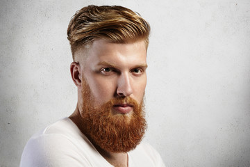 Sideways portrait of natural blond Caucasian man on white background. Bearded hipster with clean shaved temples and stylish well-trimmed moustaches looking serious and brutal with long facial hear.