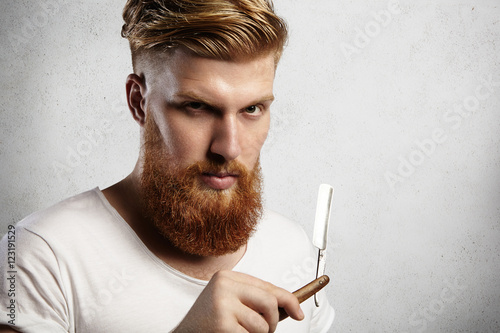 Fotografie, Obraz Headshot of attractive professional barber with thick red beard and mustache, holding razor