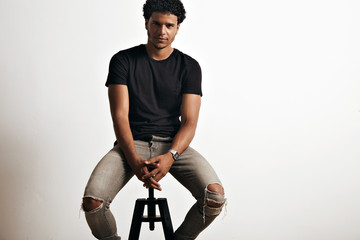 Wall Mural - Young healthy attractive black man wearing blank black t-shirt and torn grey jeans sitting on a chair in a white studio