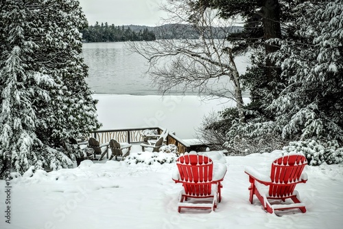 Fotografia, Obraz Pair of wooden adirondack chairs in the snow in front of the fro