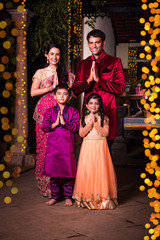 cheerful indian family welcoming on diwali night outside home with diwali lighting, indian couple and kids or people in namaskar pose welcoming guests on diwali celebration or festival night