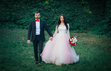  Stylish bearded groom in a tuxedo with a red butterfly and suspenders with his bride with long black hair in a pink long dress is holding a bouquet of peonies with a tied ribbon walking in green park