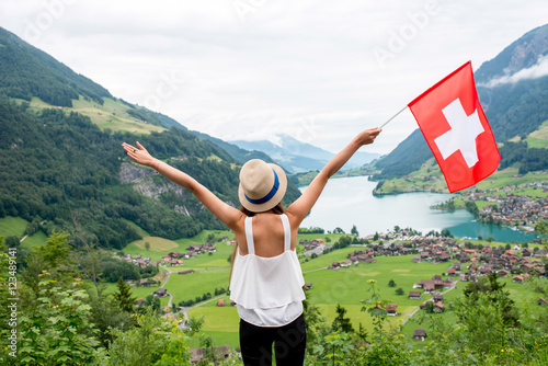 Fotografie, Obraz Young woman with swiss flag enjoying great landscape view on the valley with lake and village in Switzerland