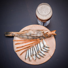 Glass of beer and assorted dried fish on a cooking sheet, traditional beer snack. Black wooden background.