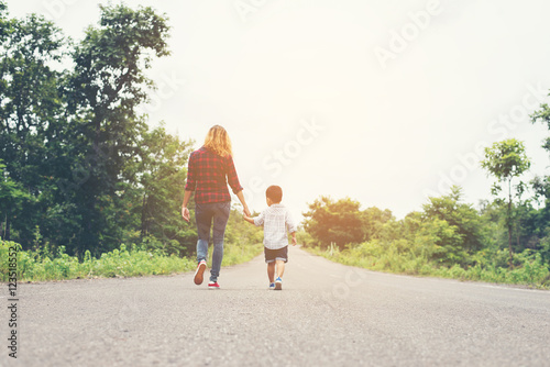 Fotografia, Obraz Mother holding a hand of his son in summer day walking on the st