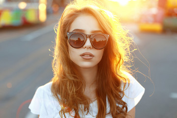 Beautiful young hipster girl in fashionable sunglasses standing at sunset 