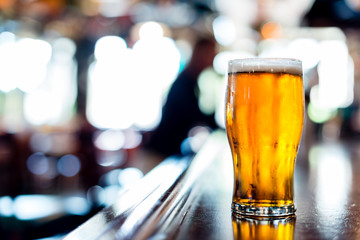 Single pint glass of glowing amber beer ale lager on long bar with blurred patron and tables in background