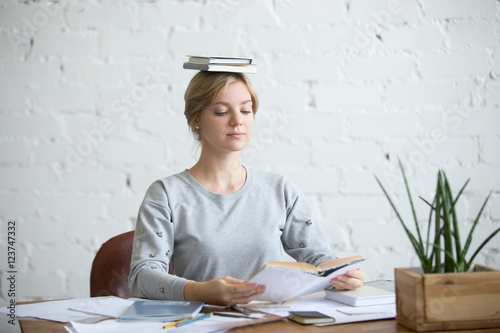 Cuadros en Lienzo Portrait of a young attractive woman at the desk with books on her head, sitting straight, reading a book