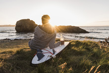 France, Bretagne, Crozon Peninsula, Woman Sitting At The Coast At Sunset With Surfboard