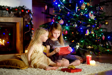 Wall Mural - Happy little sisters opening magical Christmas gift by a fireplace