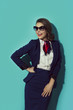 Smiling brunette stewardess wearing dark blue suit with scarf and sunglasses. Studio shot against blue wall.