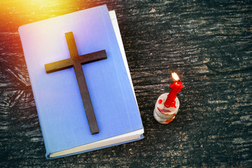 Closeup of wooden Christian cross on bible, burning candle and prayer beads on the old table. Church utensils.