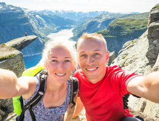 Wall Mural - Young couple doing selfie on Trolltunga. Happy man and woman enjoy beautiful lake  good weather in Norway.