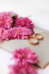 Bunch of pink chrysanthemums in an envelope and gold wedding rings. Wedding card or invitation on a white background. Season theme. Top view close-up.