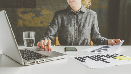 Front view of businesswoman in a gray shirt sitting at a desk and using a laptop while holding a graph. On the table there are other graphics, a smartphone and a glass of water. She uses the gadget.