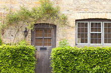 Brown Rustic Wooden Doors In An Old Traditional English Stone Honey Colored Cottage, Green Hedge In Front